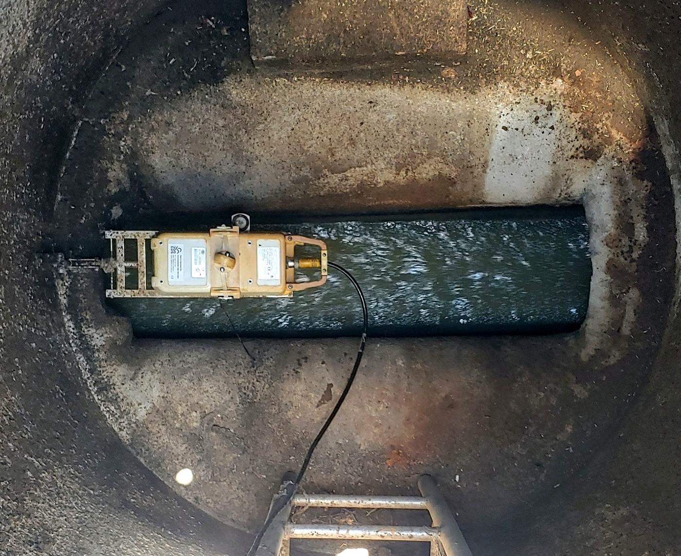 An area velocity flow meter on a mounting band at the invert of a sewer, seen from the manhole above, with wastewater flowing over it